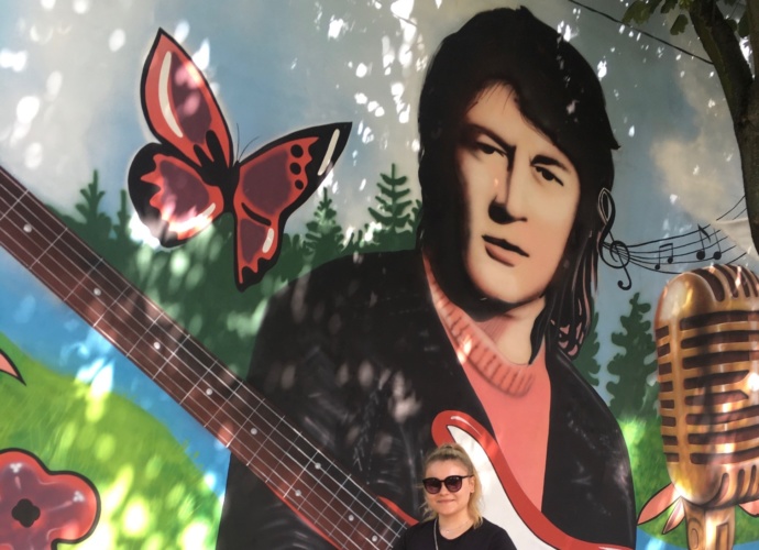 W tle fragment muralu muzycznego przy pasaży Klenczona w Pułtusku, a na nim od lewej motyl, wizerunek Krzysztofa Klenczona z gitarą oraz mikrofonem, na pierwszym planie autorka koncepcji projektu graficznego