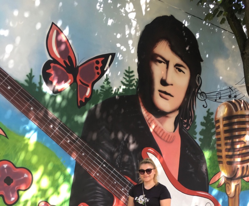 W tle fragment muralu muzycznego przy pasaży Klenczona w Pułtusku, a na nim od lewej motyl, wizerunek Krzysztofa Klenczona z gitarą oraz mikrofonem, na pierwszym planie autorka koncepcji projektu graficznego