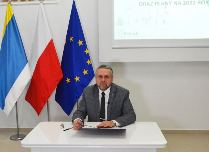 Burmistrz Wojciech Gregorczyk. Zdjęcie burmistrza siedzącego przy biurku. W tle flagi Pułtuska, Polski i Unii Europejskiej